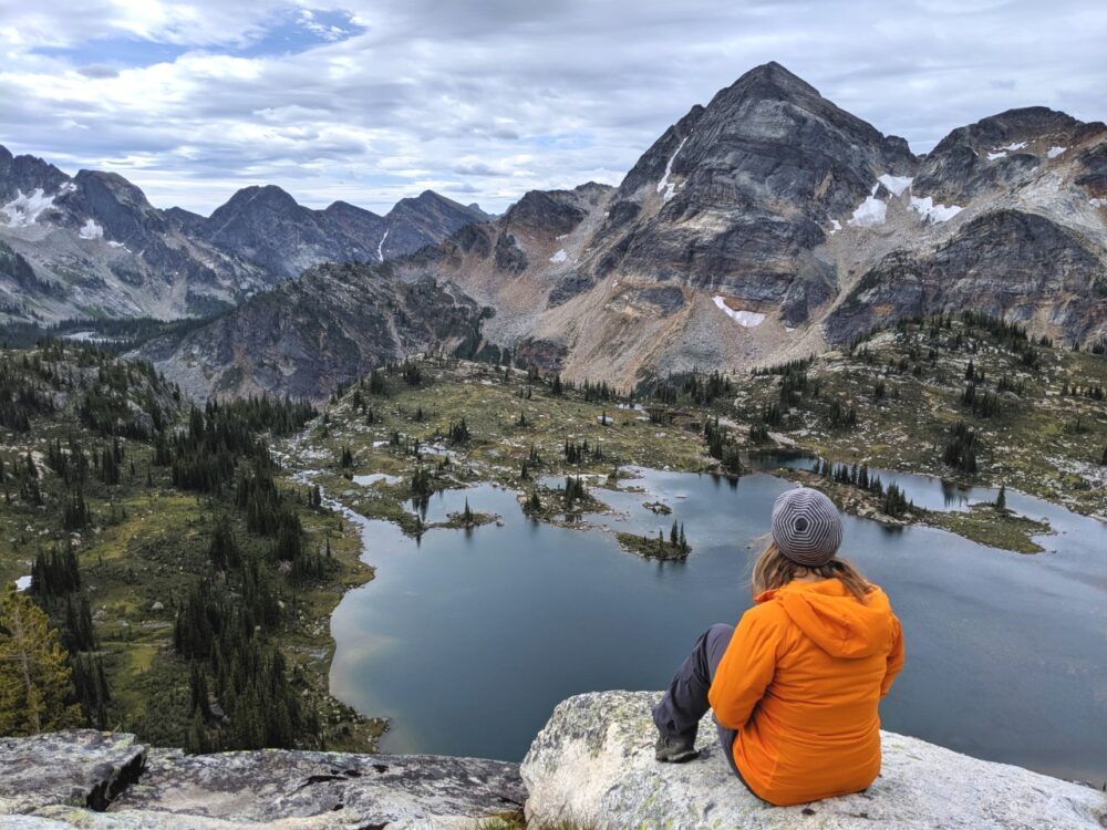 杰玛坐在岩石上，向下看下面精致的高山景观，有湖泊、草地和周围的山脉