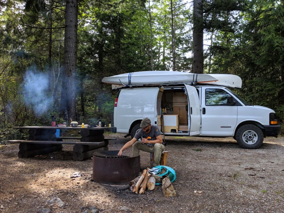 在不列颠哥伦比亚省的露营地，JR坐在野餐桌旁和白色货车前参加篝火晚会