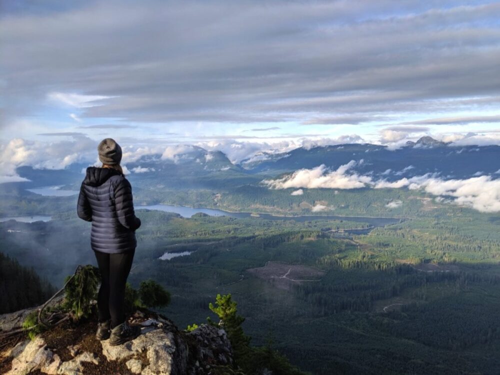 杰玛背对着镜头站在悬崖上，欣赏着阳光海岸小径上的湖泊、山脉和森林