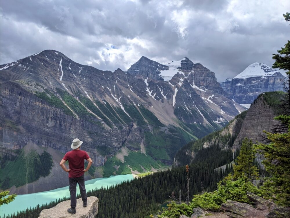 JR站在一块岩石上，双手叉腰，看着环绕着长长的、浑浊的彩色湖泊的巨大山脉的后景