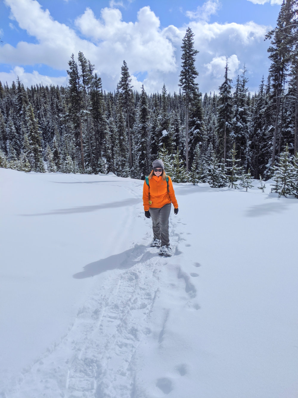 杰玛穿着橙色夹克，雪地鞋走向镜头，背景是白雪皑皑的森林