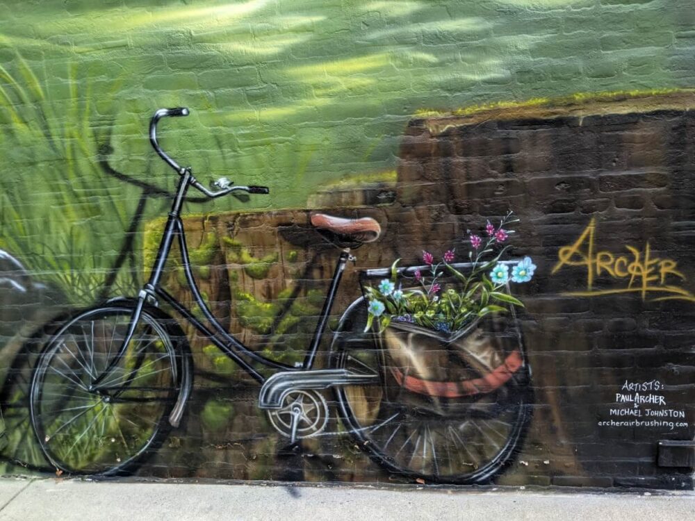 在砖建筑上画的壁画，自行车与花在袋子上的后轮胎。艺术家的名字(Archer)在右边