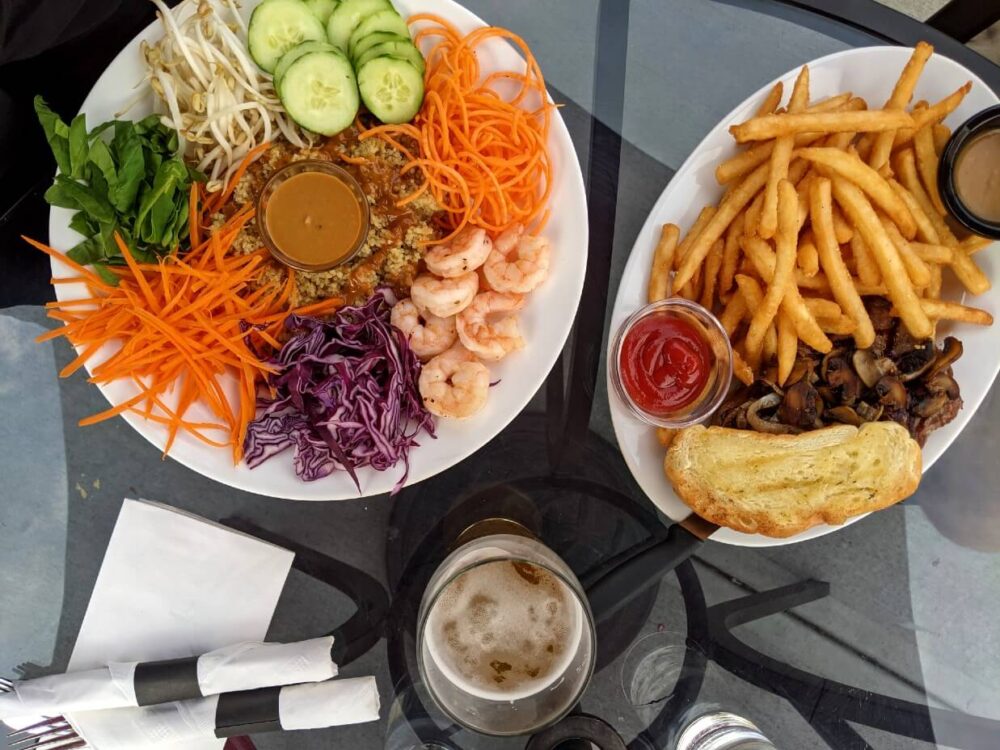 在Keg and Kettle Grill餐厅用餐的俯视图，有藜麦盘(对虾，蔬菜)和牛排配蒜蓉面包和薯条。盘子之间有啤酒和餐具