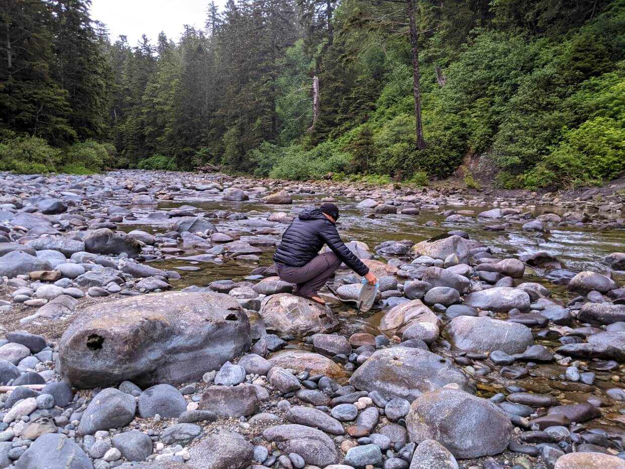 后视图JR蹲在小溪与水袋，收集水在卡玛纳河