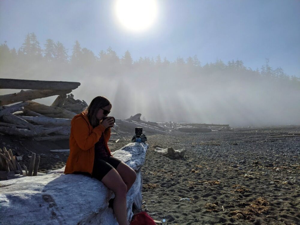 杰玛坐在沙滩上的浮木上，背景是透过雾气的阳光