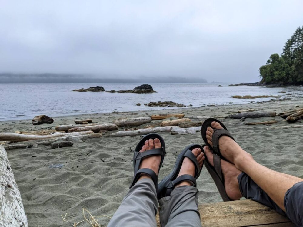 镜头后的两条腿伸展到沙滩上，两双脚都穿着深色凉鞋。