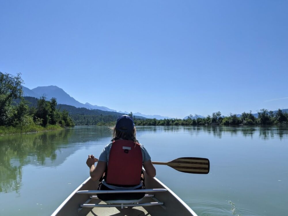 后景杰玛在独木舟上拿着桨，在平静的河流上向前看的背景山