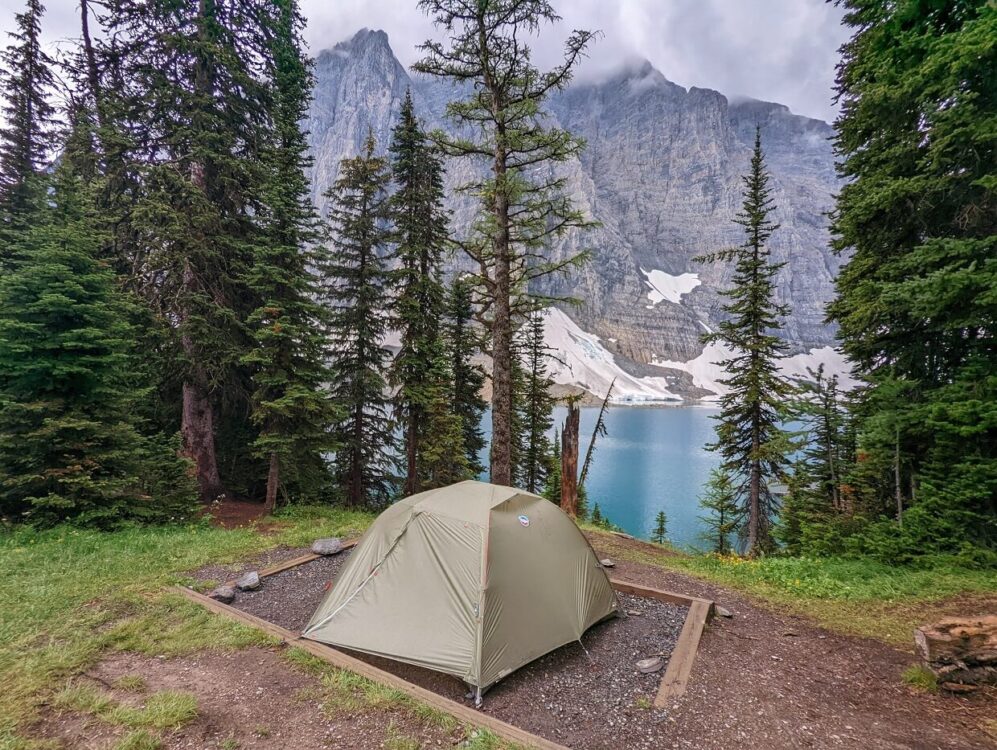 在蓝绿色浮冰湖前的砾石垫上搭起帐篷，背景是高耸的山脉
