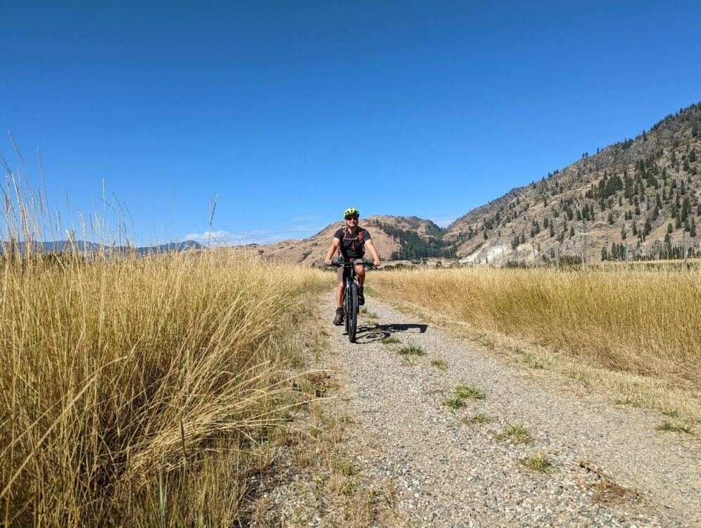 JR骑着自行车在坚硬的砾石路上向镜头走去，两边是金色的草地，背景是森林覆盖的小山