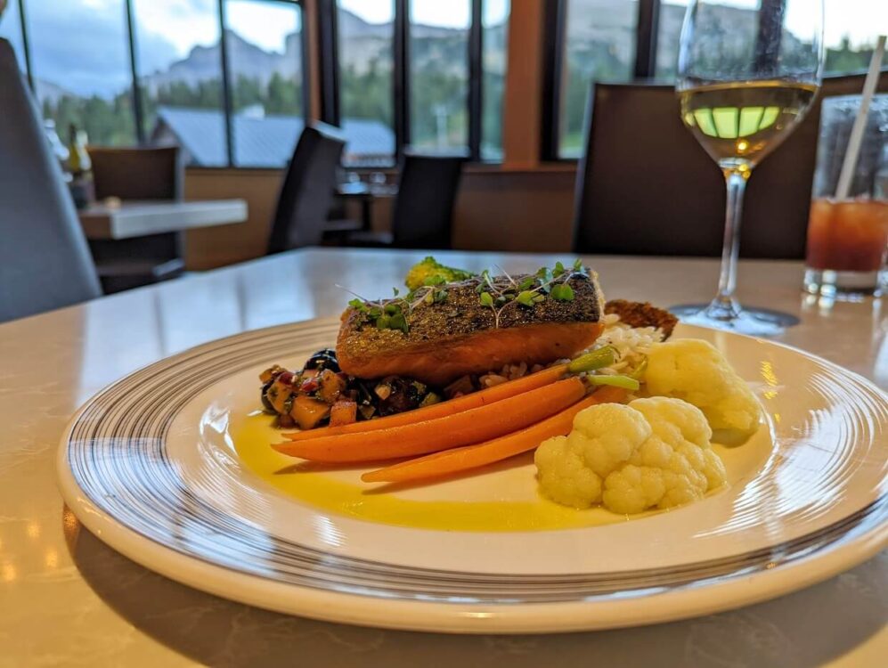 烟囱角餐厅的三文鱼菜与花椰菜，胡萝卜和莎莎酱。在白色的盘子旁边有一杯白葡萄酒。背景中可以看到窗户，远处是山