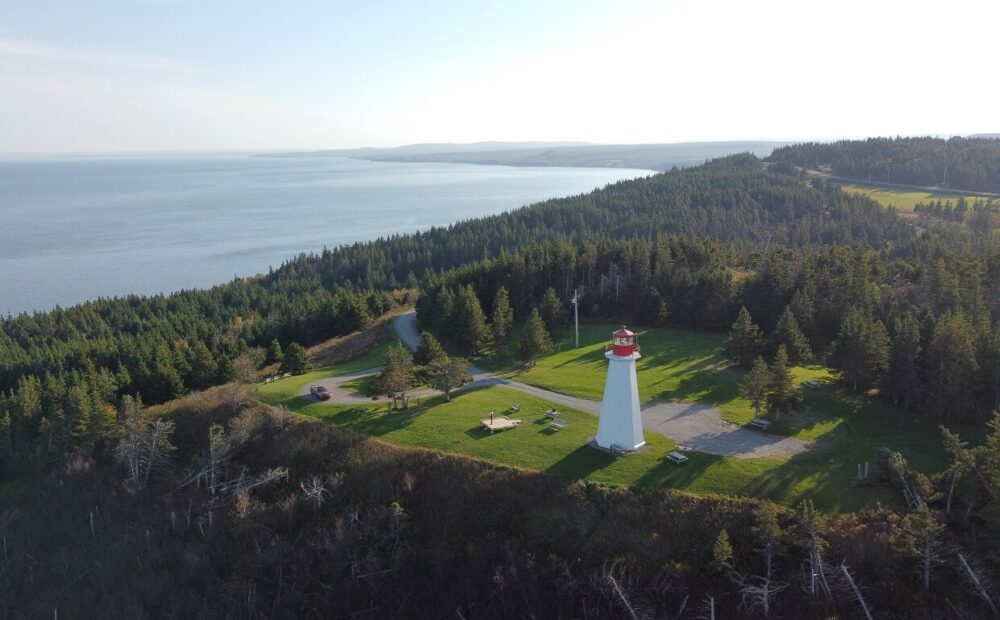 安提戈尼什附近的乔治角灯塔无人机视图，红色和白色的灯塔位于悬崖边缘，周围是森林。左边可以看到海洋