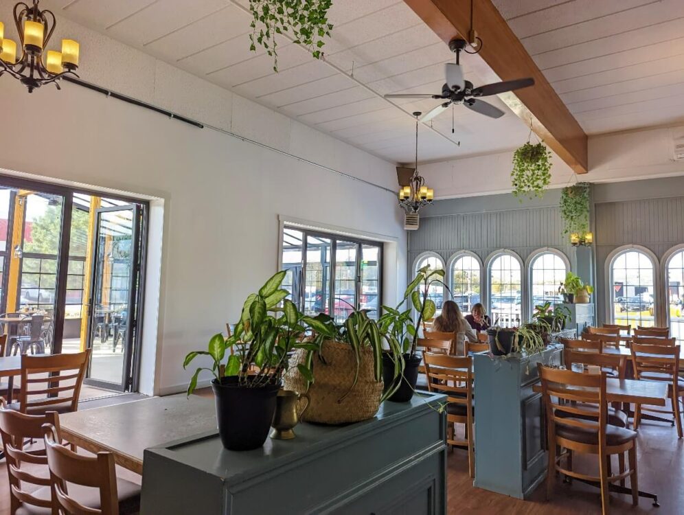 透过Justamere Cafe and Bakery明亮通风的用餐空间，高高的天花板、许多植物、木桌、木地板和白墙。有一张桌子被占用了